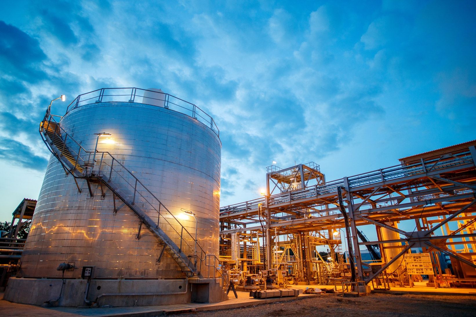Insulated storage tank with an external stairway beside a lit process pipe rack at dusk