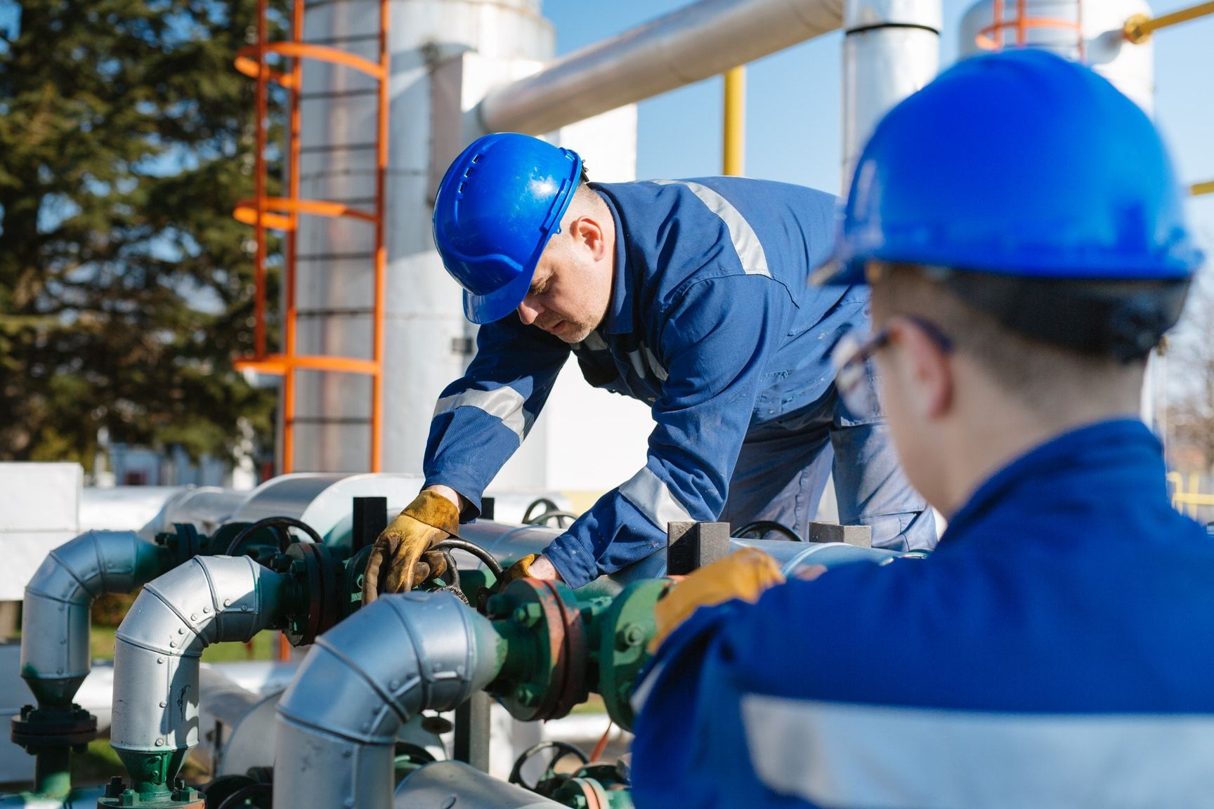 Two technicians in blue coveralls and hard hats operating a valve handwheel on process piping
